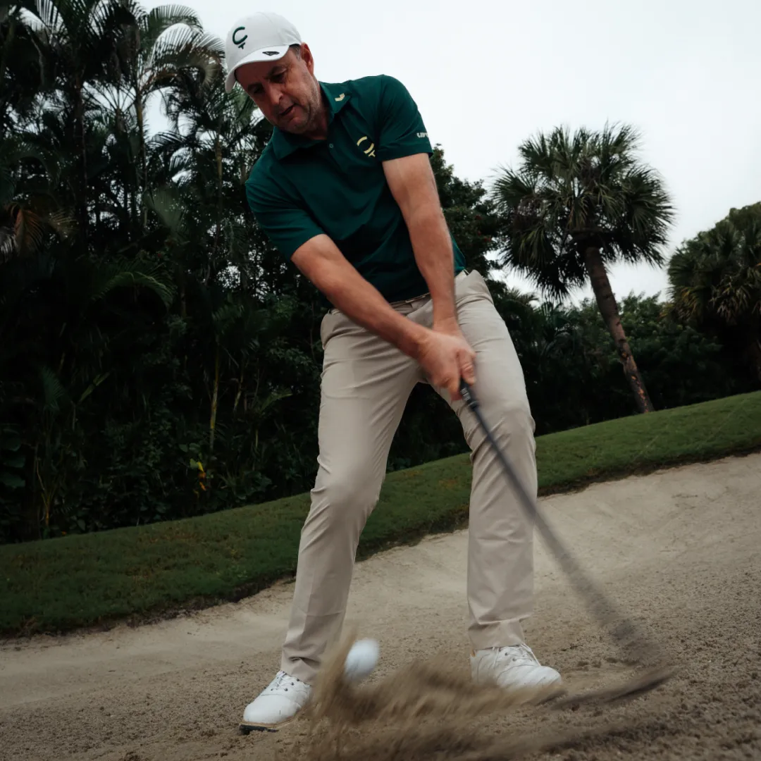 Golfer in a green shirt and white cap swinging a club, hitting a ball from a sandy bunker with trees in the background.