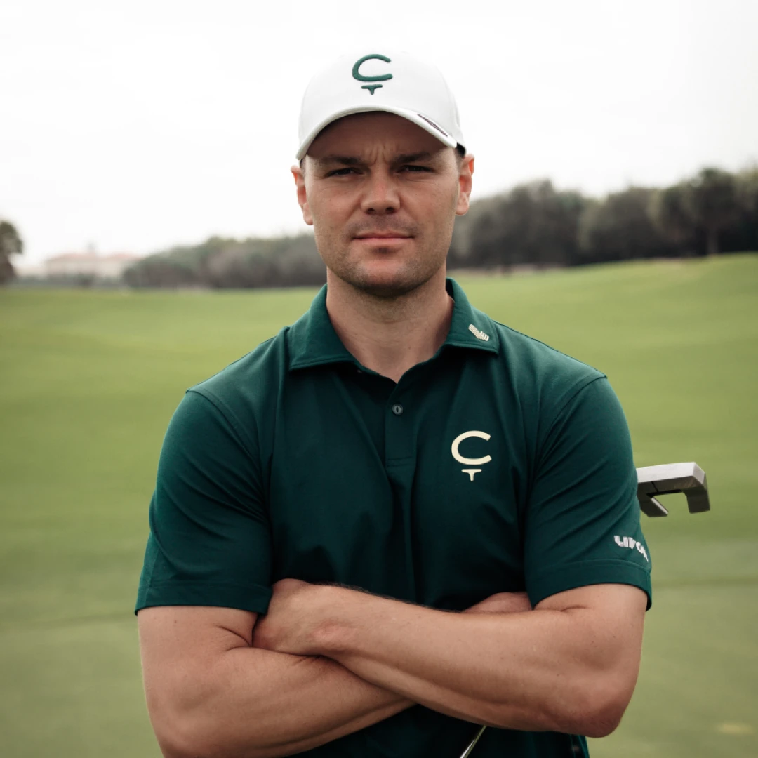 Golfer in a green shirt and white cap stands with arms crossed on a golf course, holding a club in his left hand.
