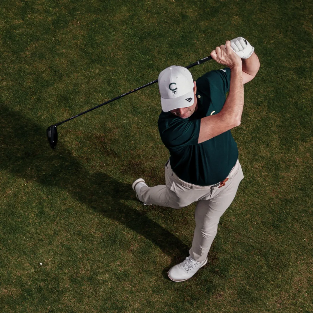 Golfer in a cap swings a club on a green golf course, wearing a dark green shirt and beige pants, viewed from above.