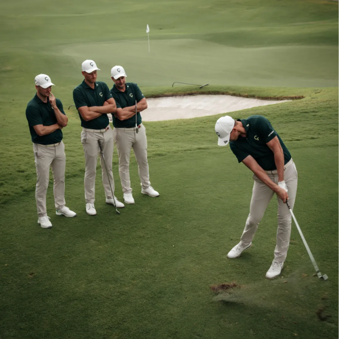 Four golfers in matching attire on a course, with one swinging a club while the others observe. A sand trap is visible in the background.