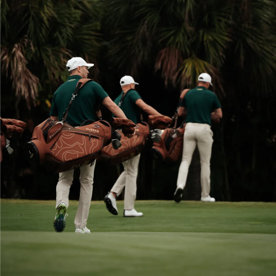 Three golfers in green shirts and white caps walk across a golf course carrying brown golf bags, with trees in the background.